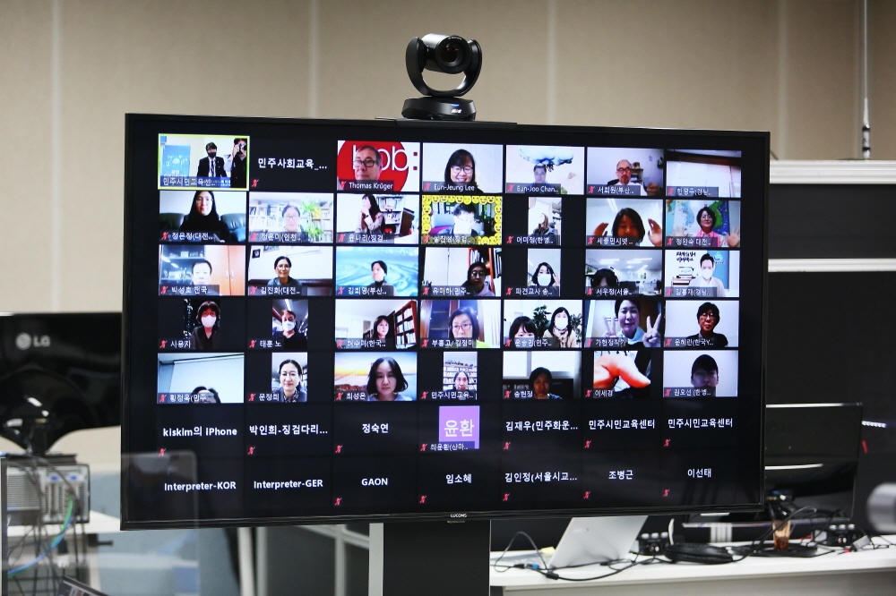 회의실에 연결된 텔레비전에 ZOOM 화상회의로 연결되어 보이는 참여자들의 사진